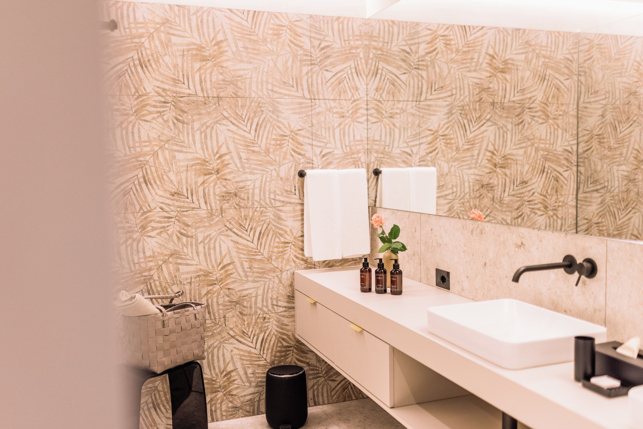 Un baño con papel tapiz estampado con hojas, un lavamanos blanco, un cesto de basura negro y un perchero con dos toallas.