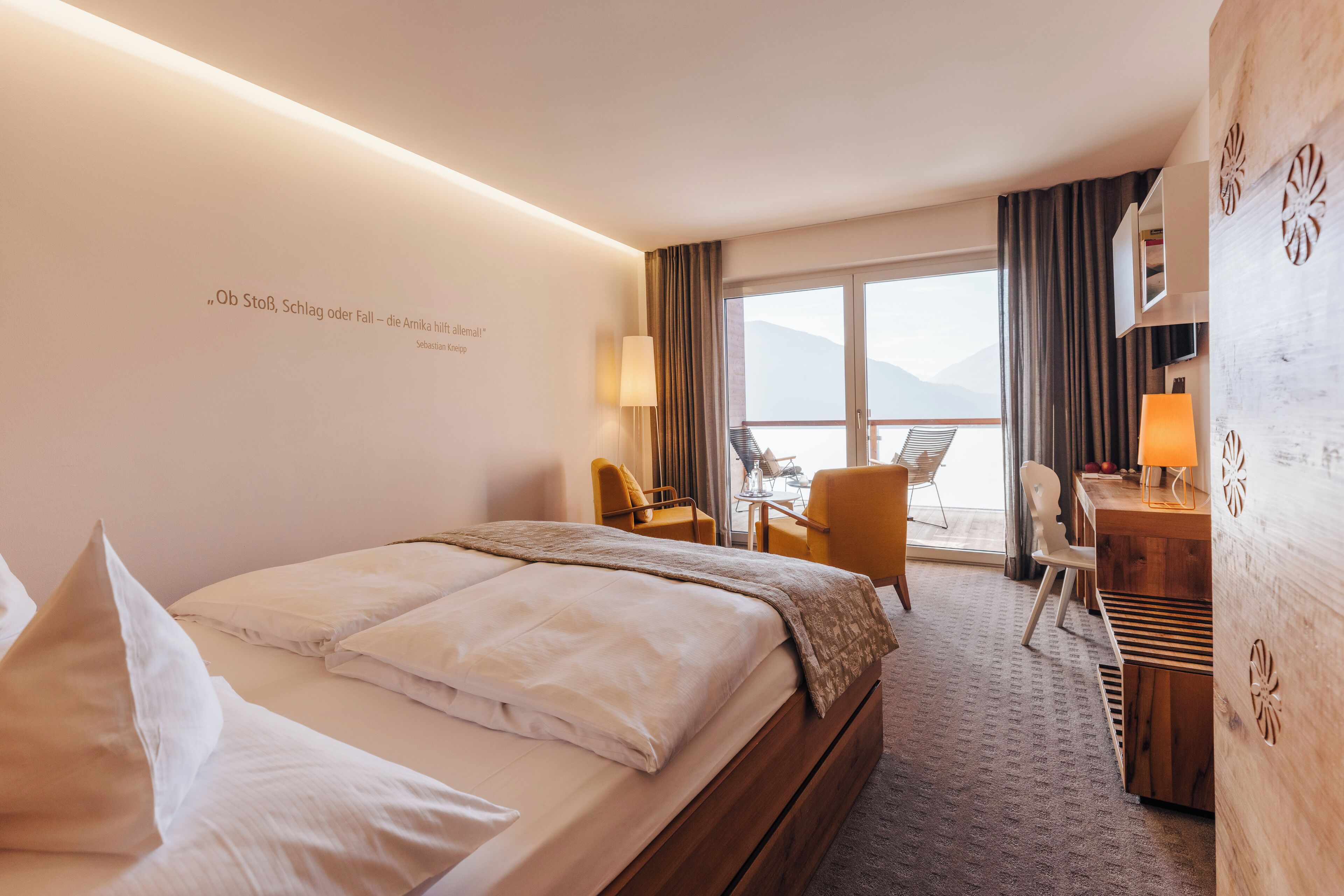 A hotel room with a bed, a desk, a chair, and a view of the mountains.