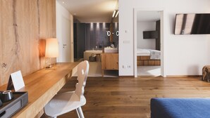 A modern hotel room with a wooden desk, a chair, a bed, a TV, and a bathroom with a sink and mirror. - Hotel Hohenwart (Schenna)