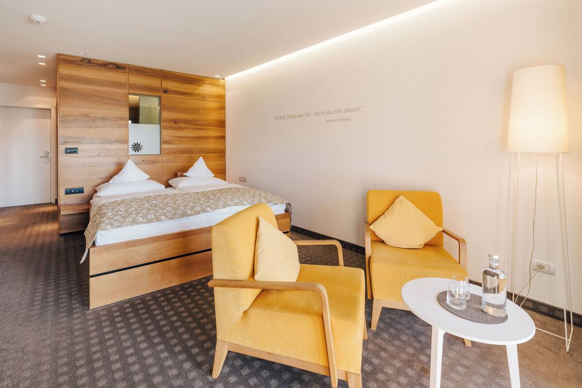 A hotel room with a bed, two yellow chairs, a small white table, and a lamp.