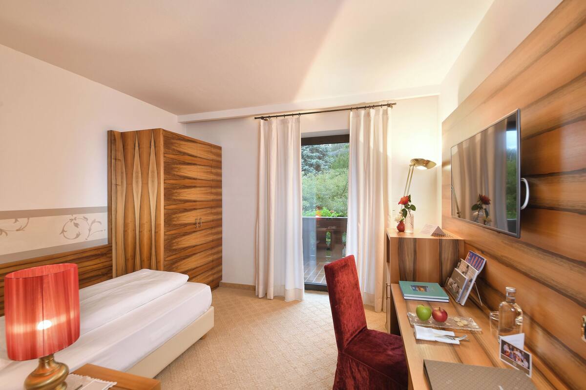 A hotel room with a bed, a desk with a lamp, a chair, a TV, and a view of greenery outside.
