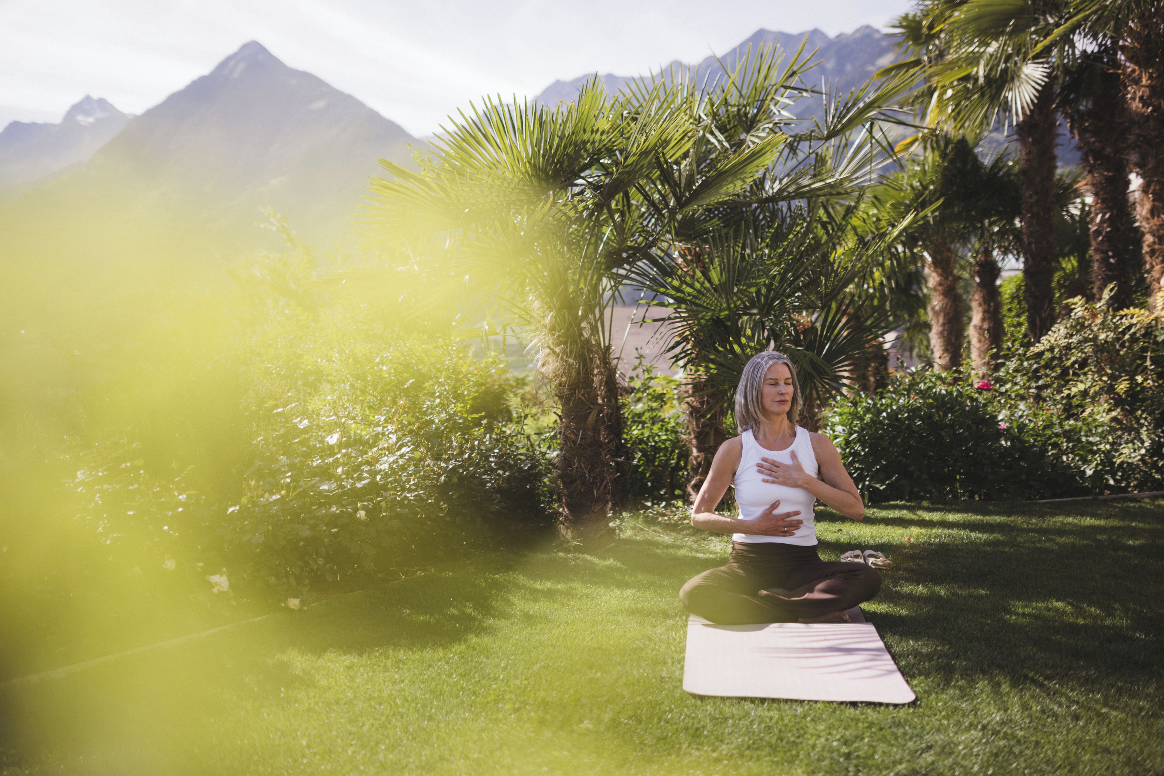 A person meditating on a yoga mat in a garden with mountains in the background.