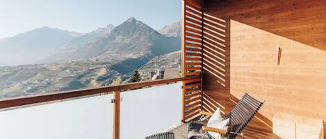 A wooden balcony with a view of mountains, a table with a bottle, and two chairs.