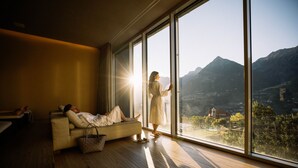 A room with a large window, a sofa, and a person standing by the window. - Hotel Hohenwart (Schenna)