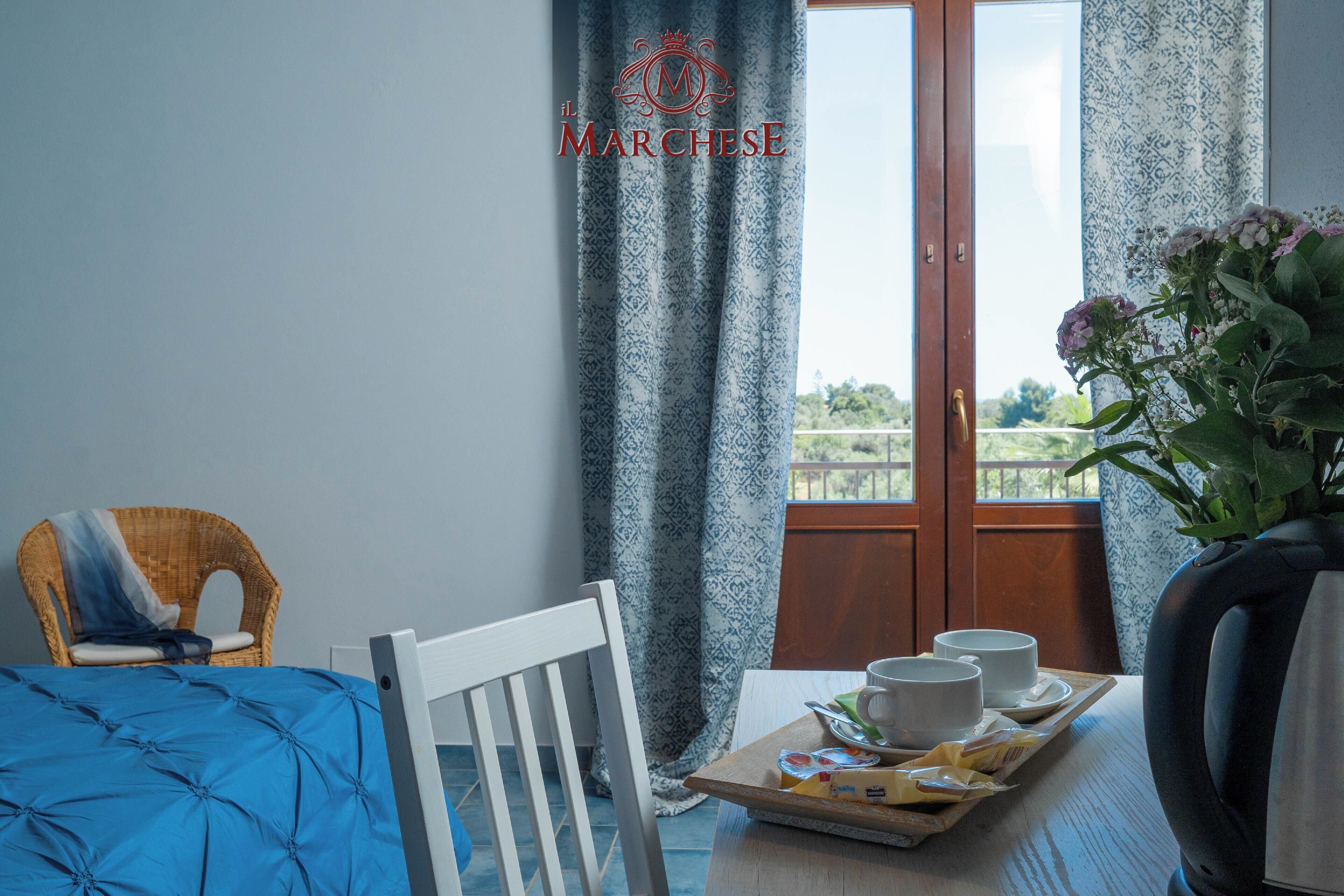 A hotel room with a bed, a chair, a table with a tray of items, and a view of greenery through a window.