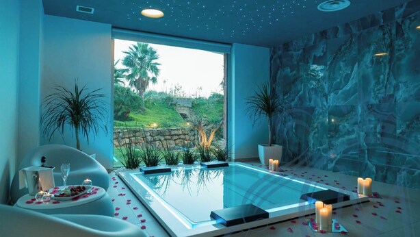 A spa room with a glass-enclosed hot tub, a dining area with a table set for two, and a view of a garden with palm trees and a stone wall. - Il Marchese Hotel & Spa (Sciacca)