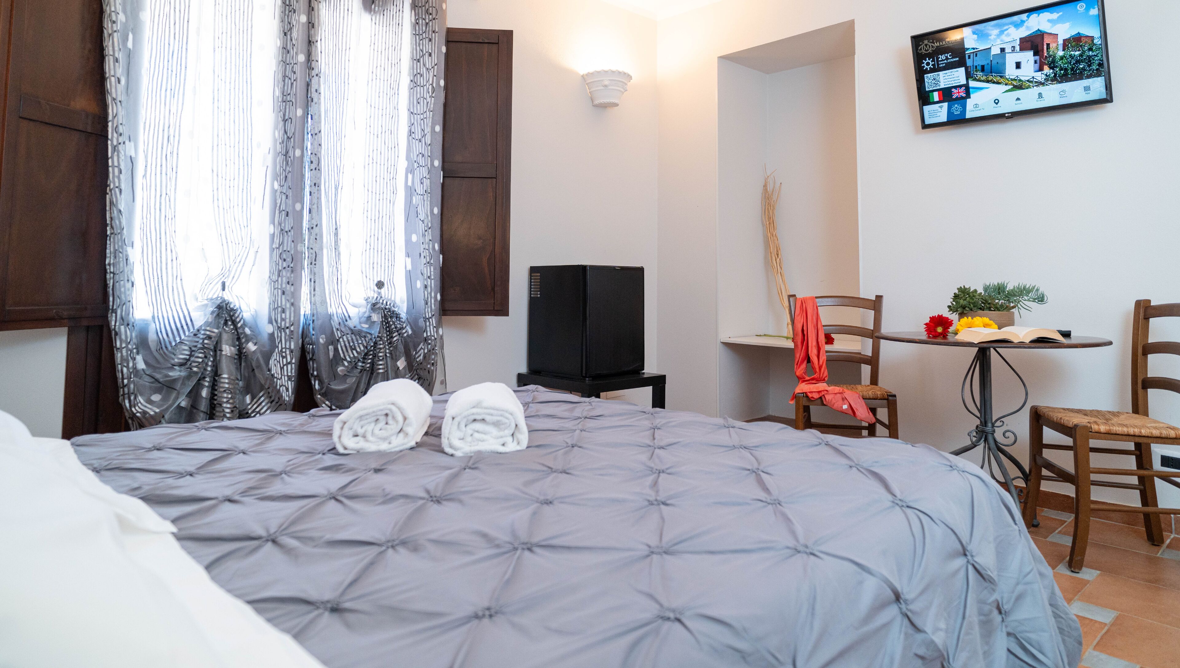 Una camera d'albergo con un letto, una televisione su un mobile, una sedia, un tavolino con una pianta e una finestra con tende.