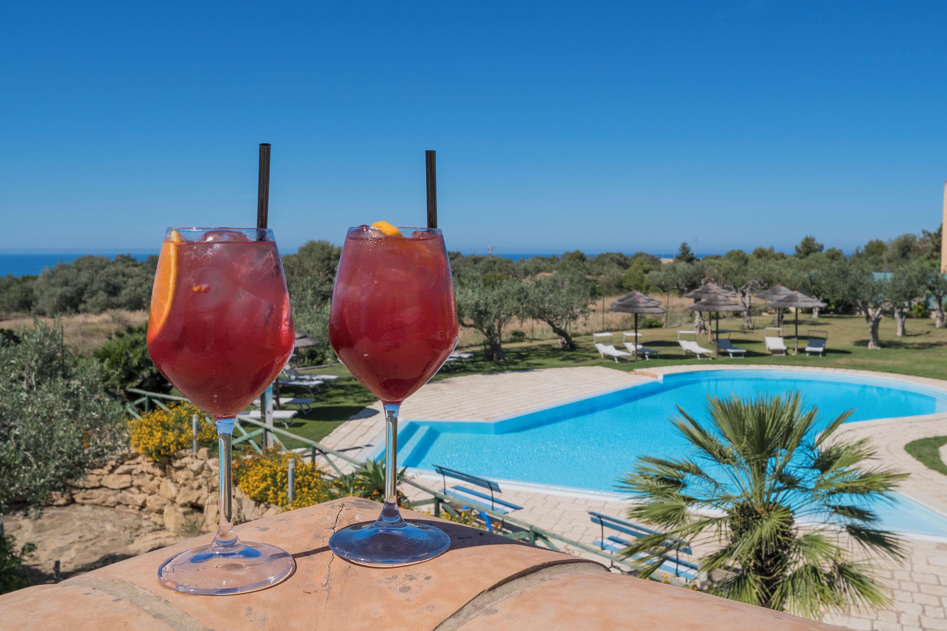 Zwei Gläser mit rotem Getränk und Trinkhalmen auf einem Tisch, mit Blick auf einen Swimmingpool und das Meer.