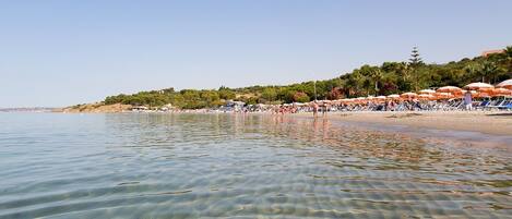 Ein Strand mit klarem, seichten Wasser, Sonnenschirmen und Menschen.