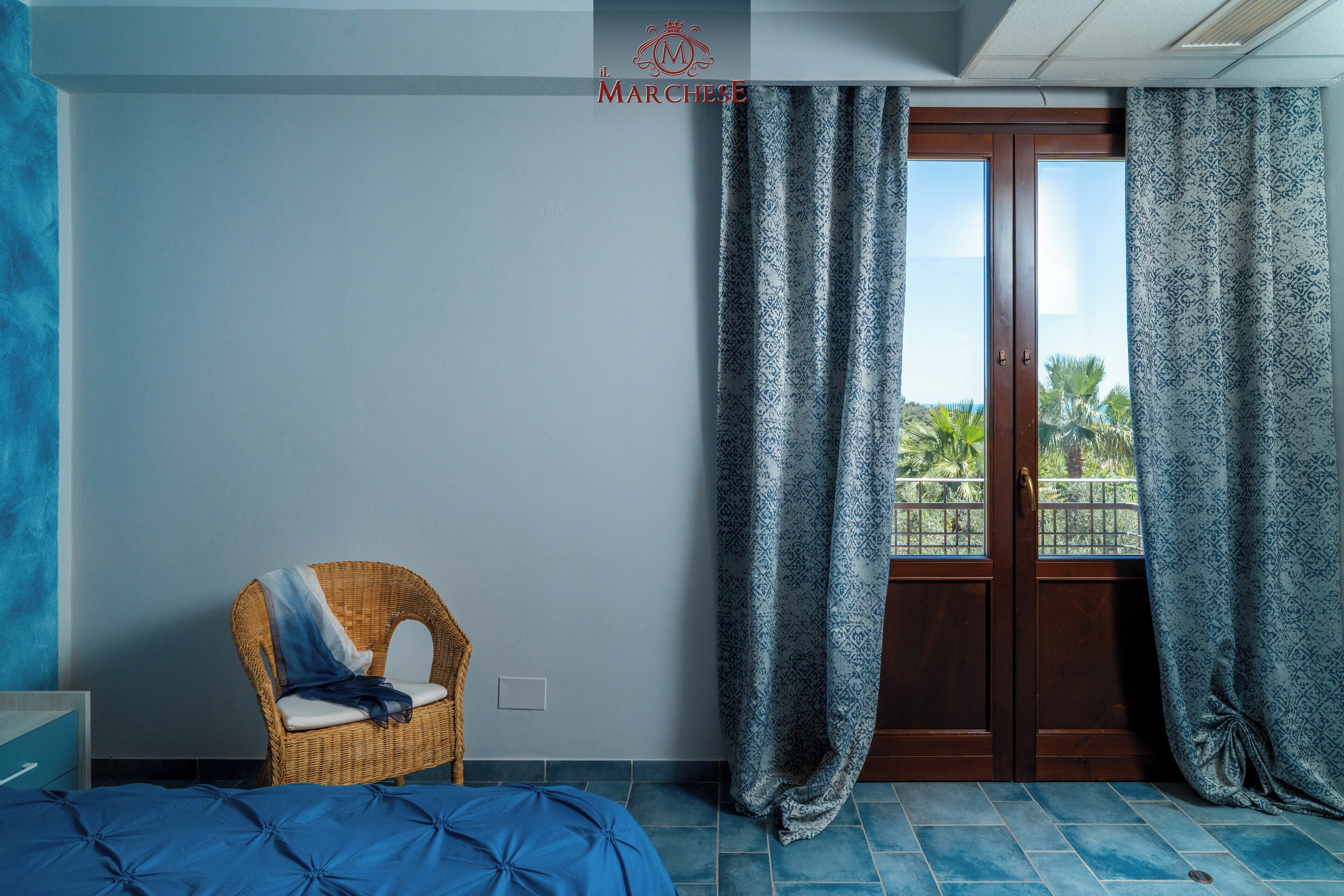 Una habitación con cama, una silla de mimbre, una ventana con cortinas estampadas y vistas a palmeras.