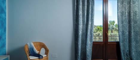 A room with a bed, a wicker chair, a window with patterned curtains, and a view of palm trees.