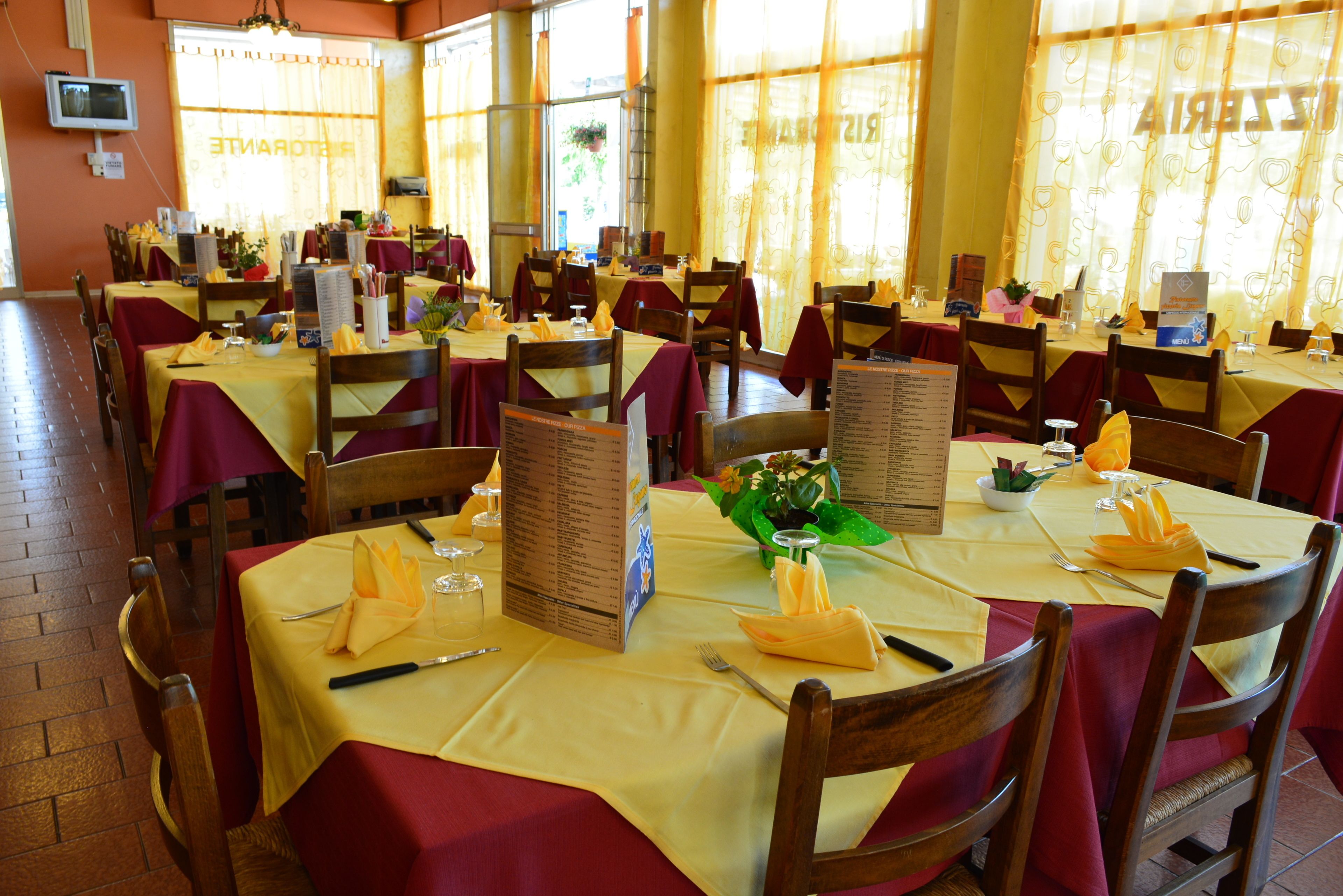 A restaurant with multiple tables set with yellow tablecloths and red napkins, wooden chairs, and menus.