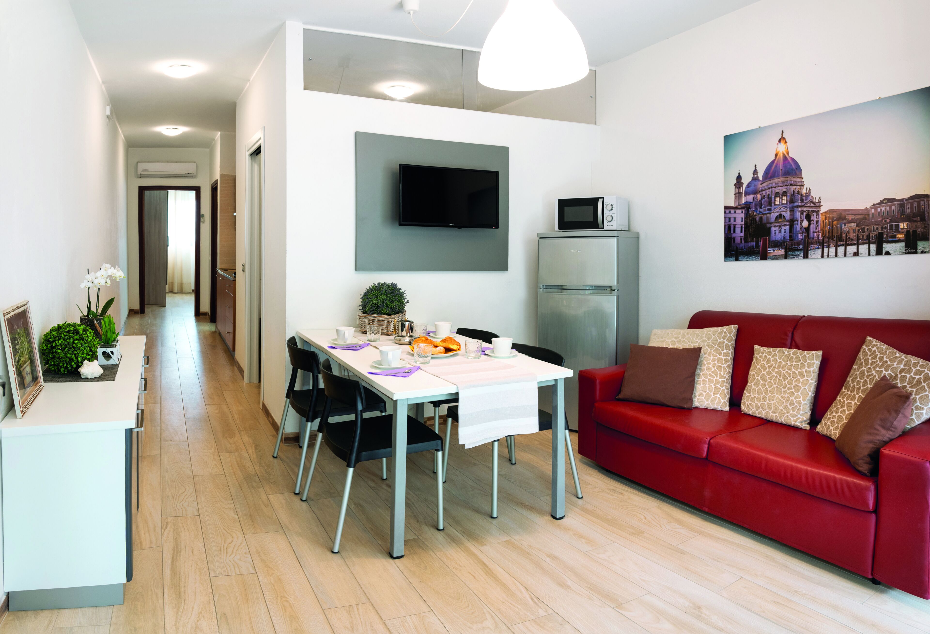 Een moderne woonkamer met een rode bank, een eettafel met stoelen, een aan de muur bevestigde televisie en een koelkast.