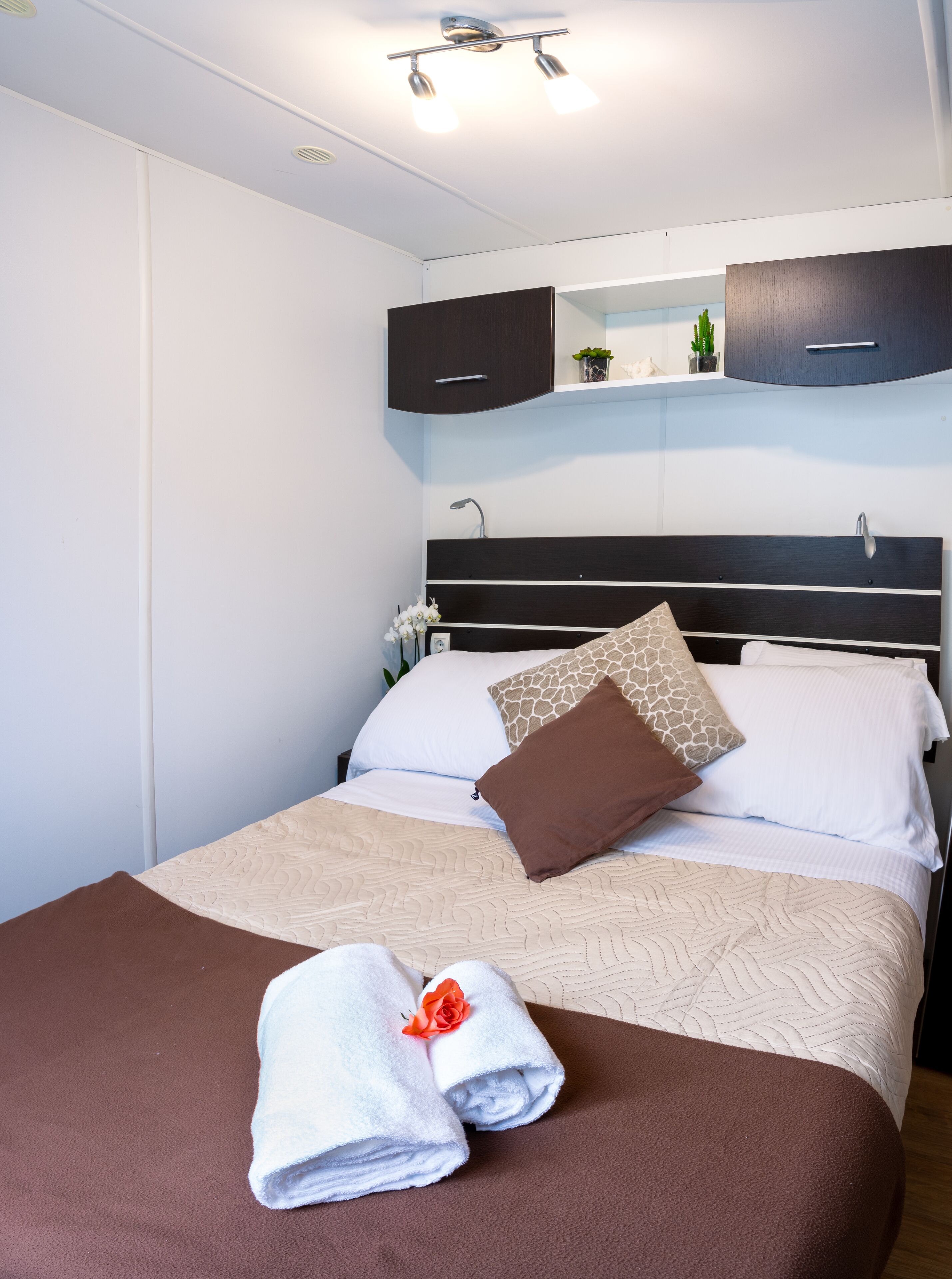 Ein ordentlich bezogenes Bett mit einer braunen und weißen Steudecke, zwei Handtücher mit einer Rosenstickerei und ein Nachttisch mit einer Lampe.