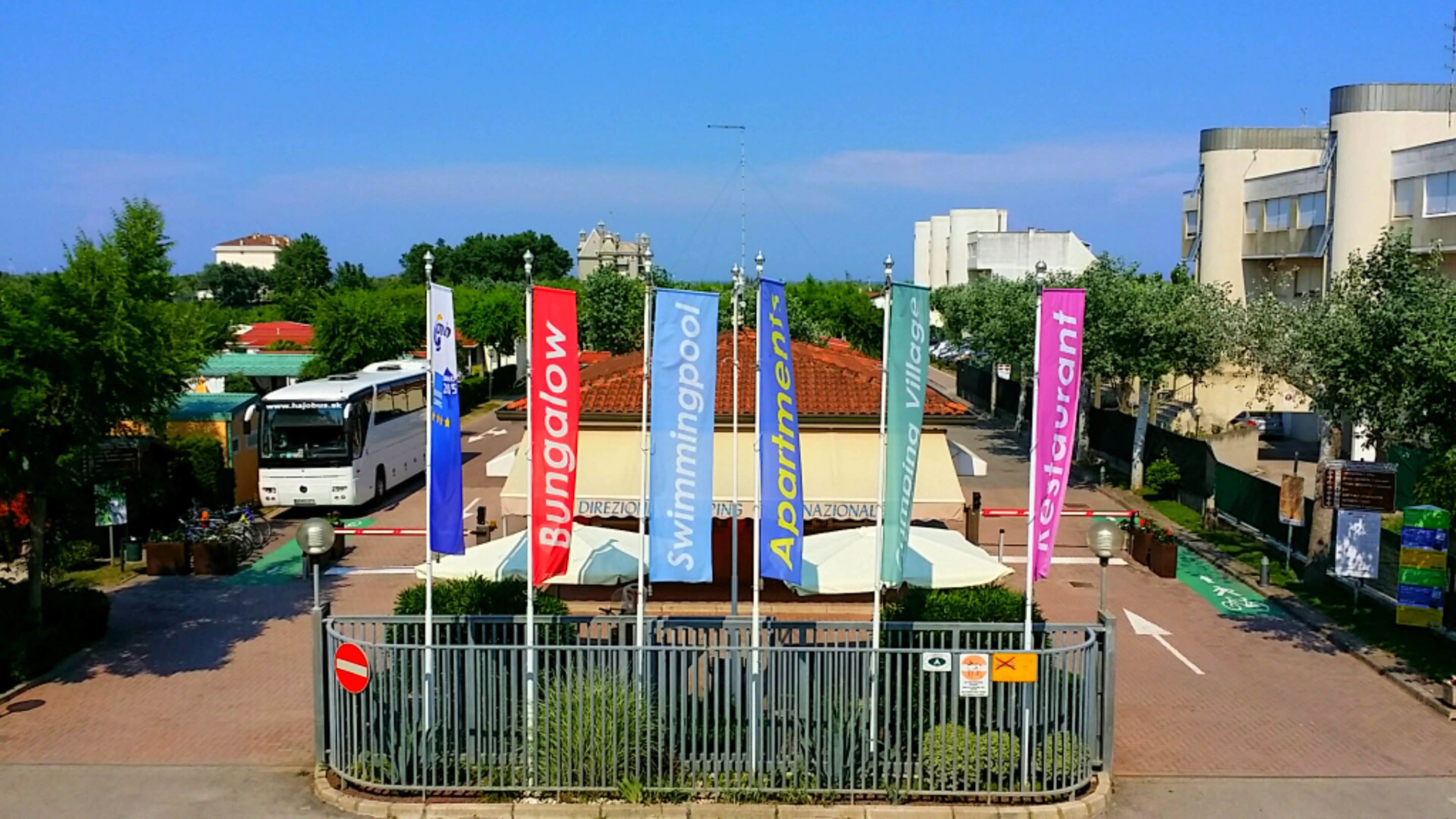 Une entrée fermée par un portail, avec des drapeaux représentant un hôtel, un autobus et un restaurant en arrière-plan.
