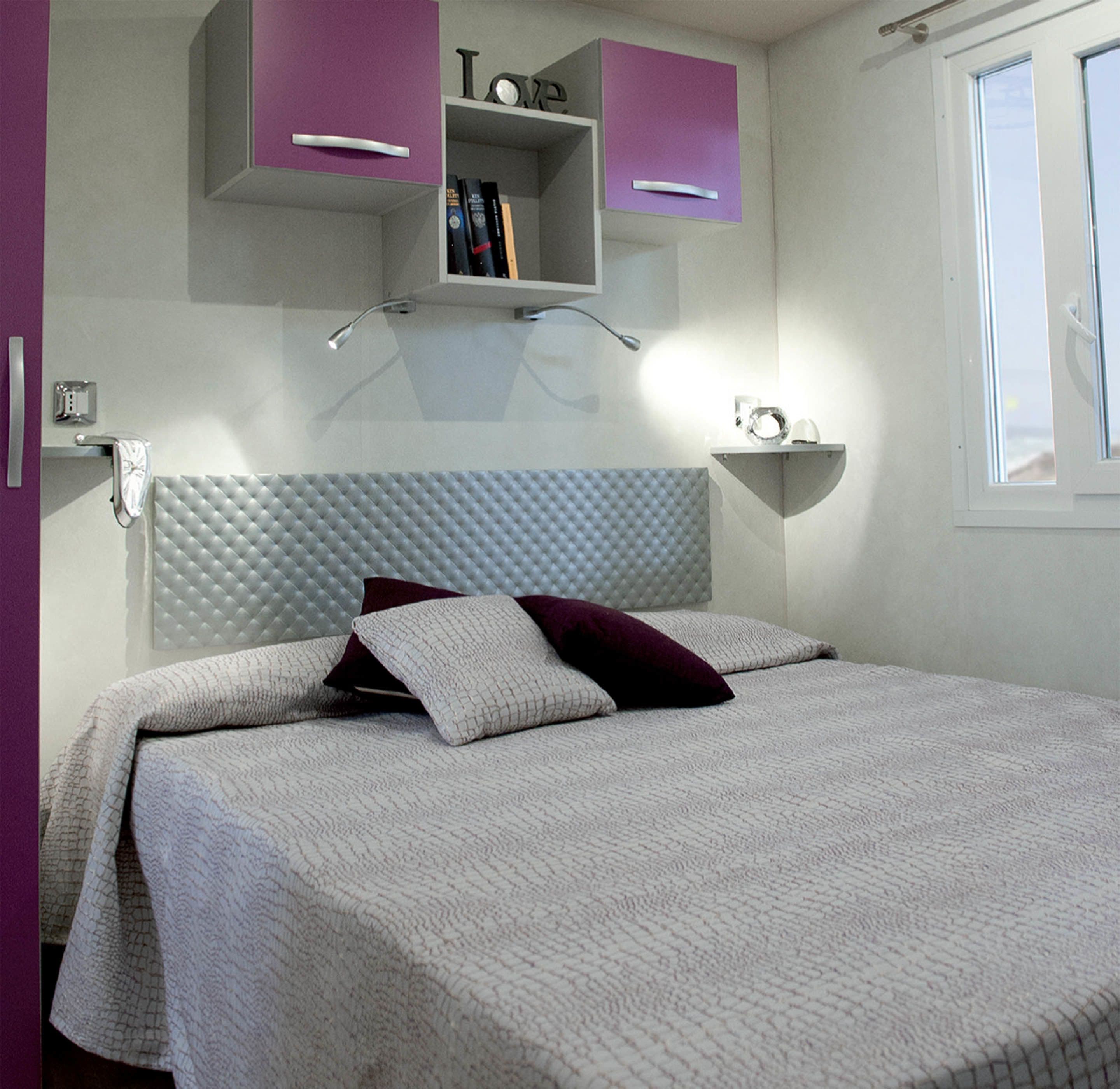 A bedroom with a tufted headboard, a bed with white bedding and dark pillows, a purple cabinet above the bed, and a small shelf with books.