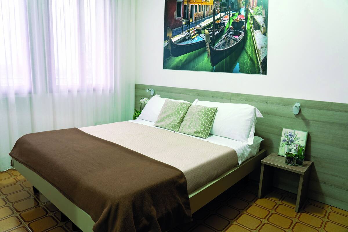A neatly made bed with a brown blanket, a small bedside table with a plant, and a painting of a canal scene on the wall.