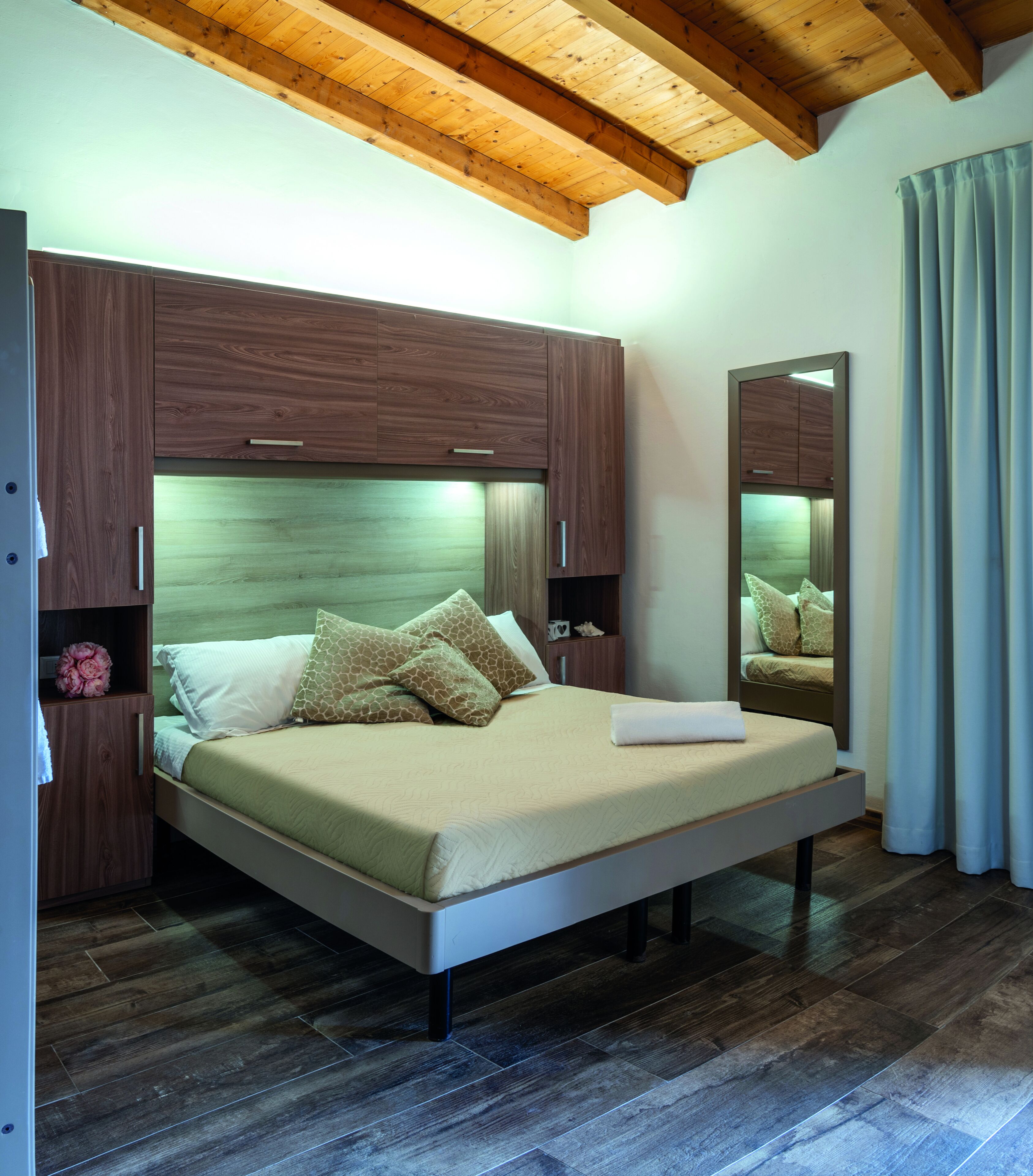 A bedroom with a wooden ceiling, a bed with a beige mattress and pillows, a nightstand, a mirror, and a window with curtains.