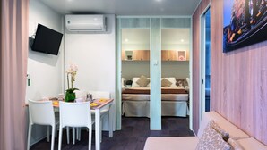 A compact hotel room with a dining area, a bed with two pillows, a mounted TV, and an air conditioning unit. - Camping Village Internazionale (Chioggia)