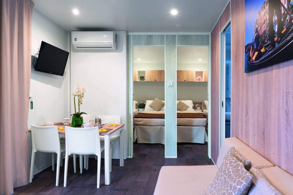 A compact hotel room with a dining area, a bed with two pillows, a mounted TV, and an air conditioning unit.
