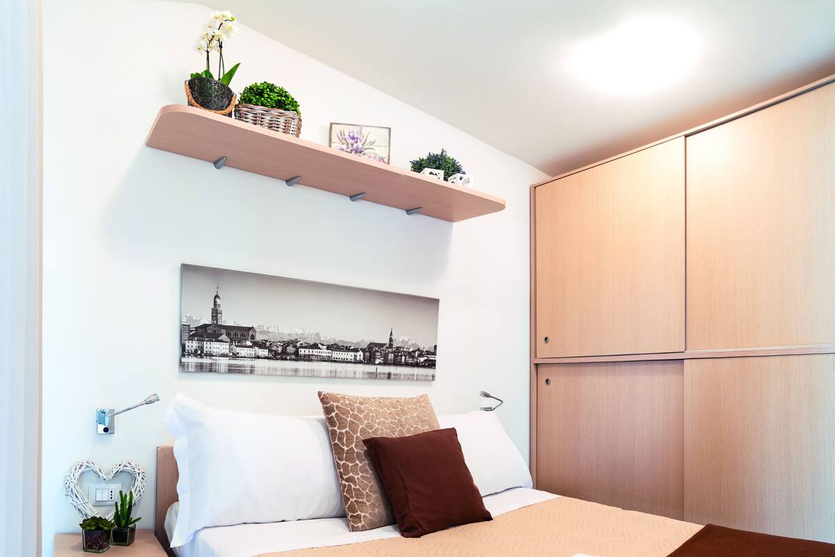 A neatly arranged bedroom with a bed, a nightstand, a wardrobe, and a wall-mounted shelf with plants and a picture.
