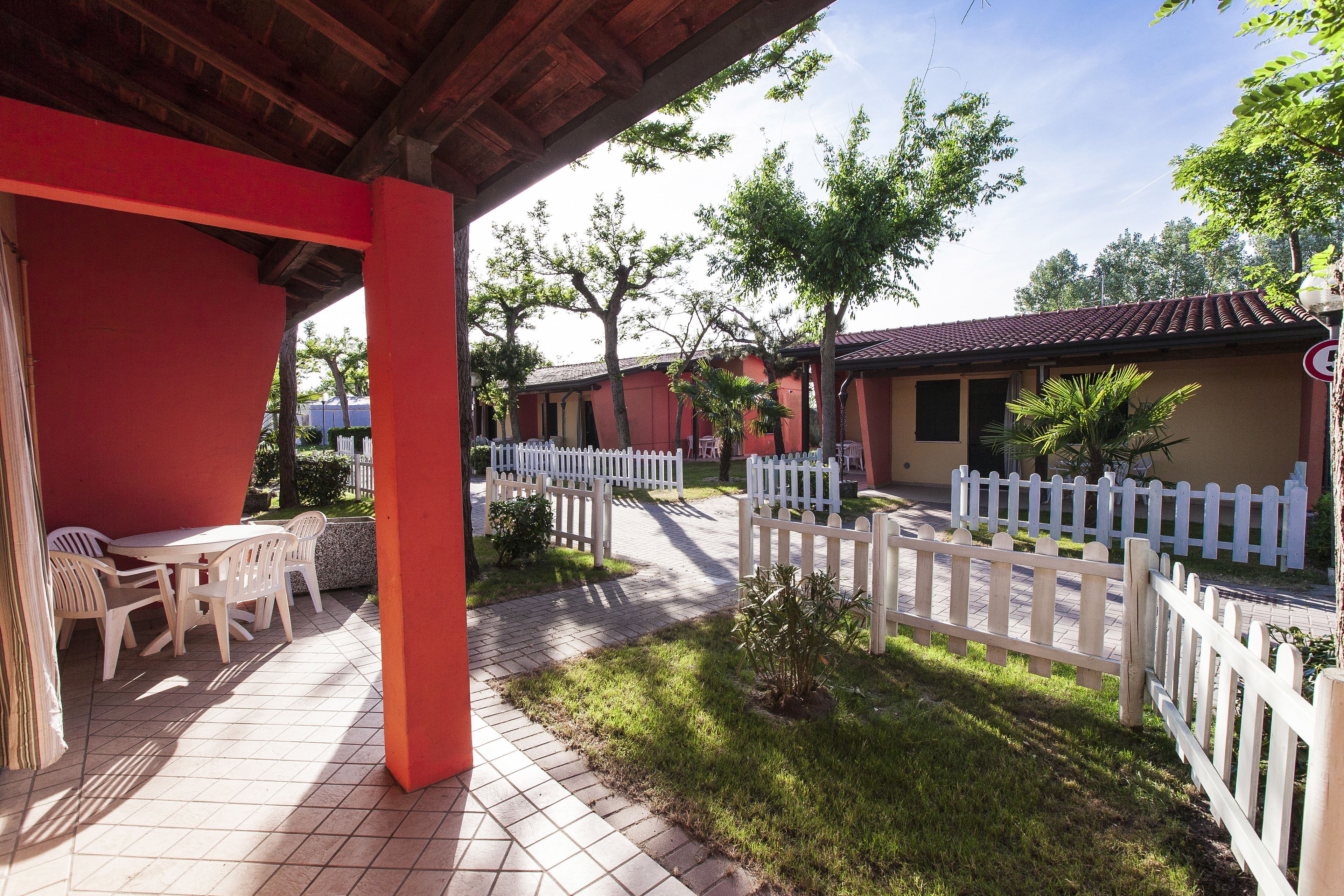A covered outdoor area with a table and chairs, a white picket fence, and a pathway leading to a building with a red roof.