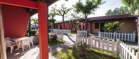 A covered outdoor area with a table and chairs, a white picket fence, and a pathway leading to a building with a red roof.