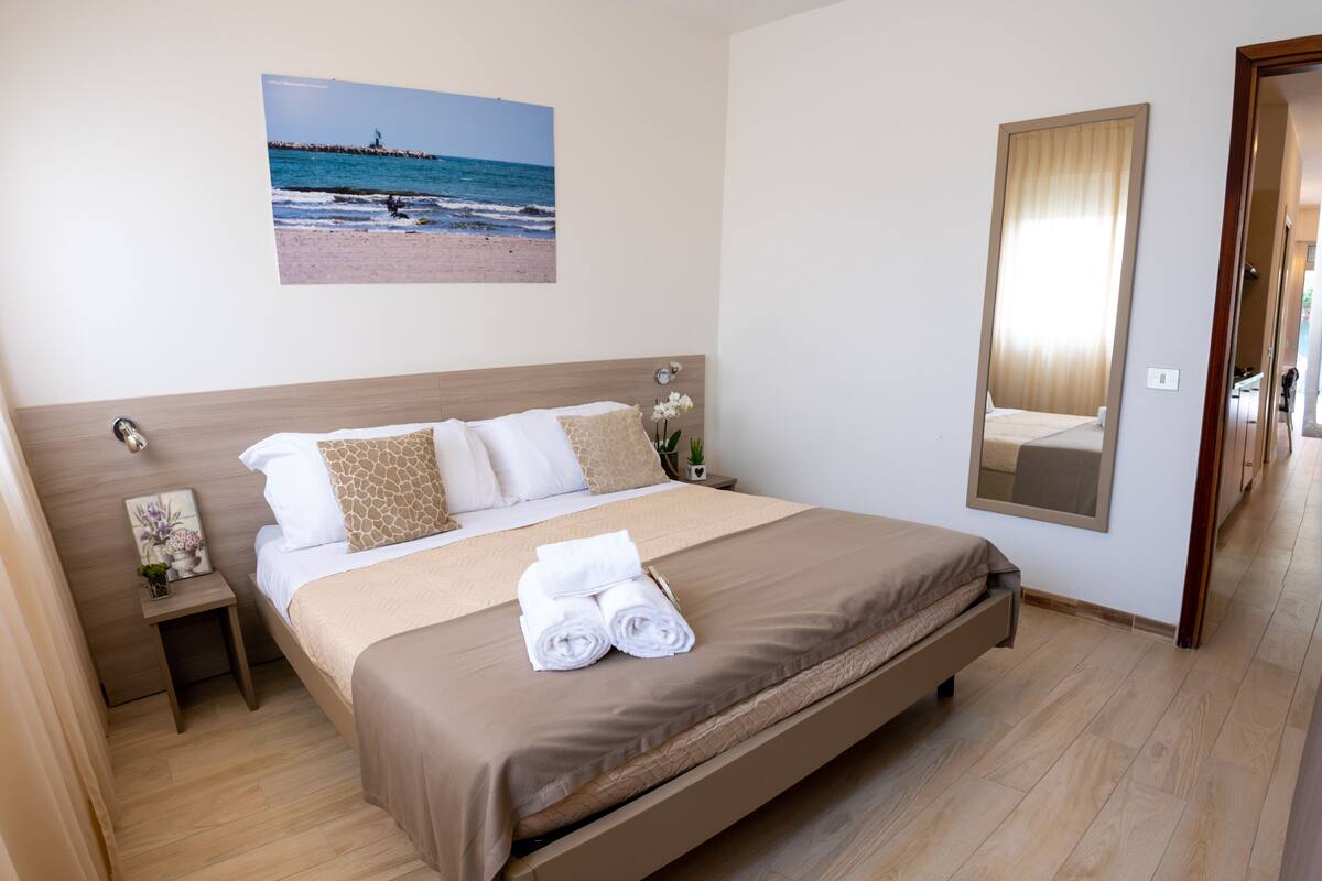 A hotel room with a large bed, a nightstand with a lamp, a framed picture on the wall, and a mirror.