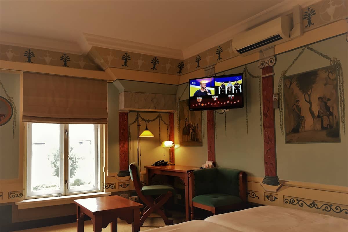 A hotel room with a bed, a desk with a lamp, a chair, a television, and a window with a view of trees.