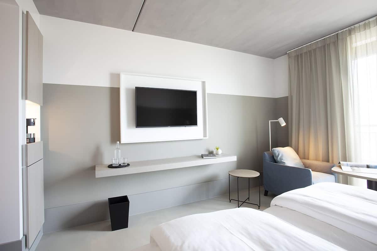 Una habitación de hotel moderna con una cama grande, un sofá, un televisor montado en la pared, una mesita auxiliar y una lámpara.