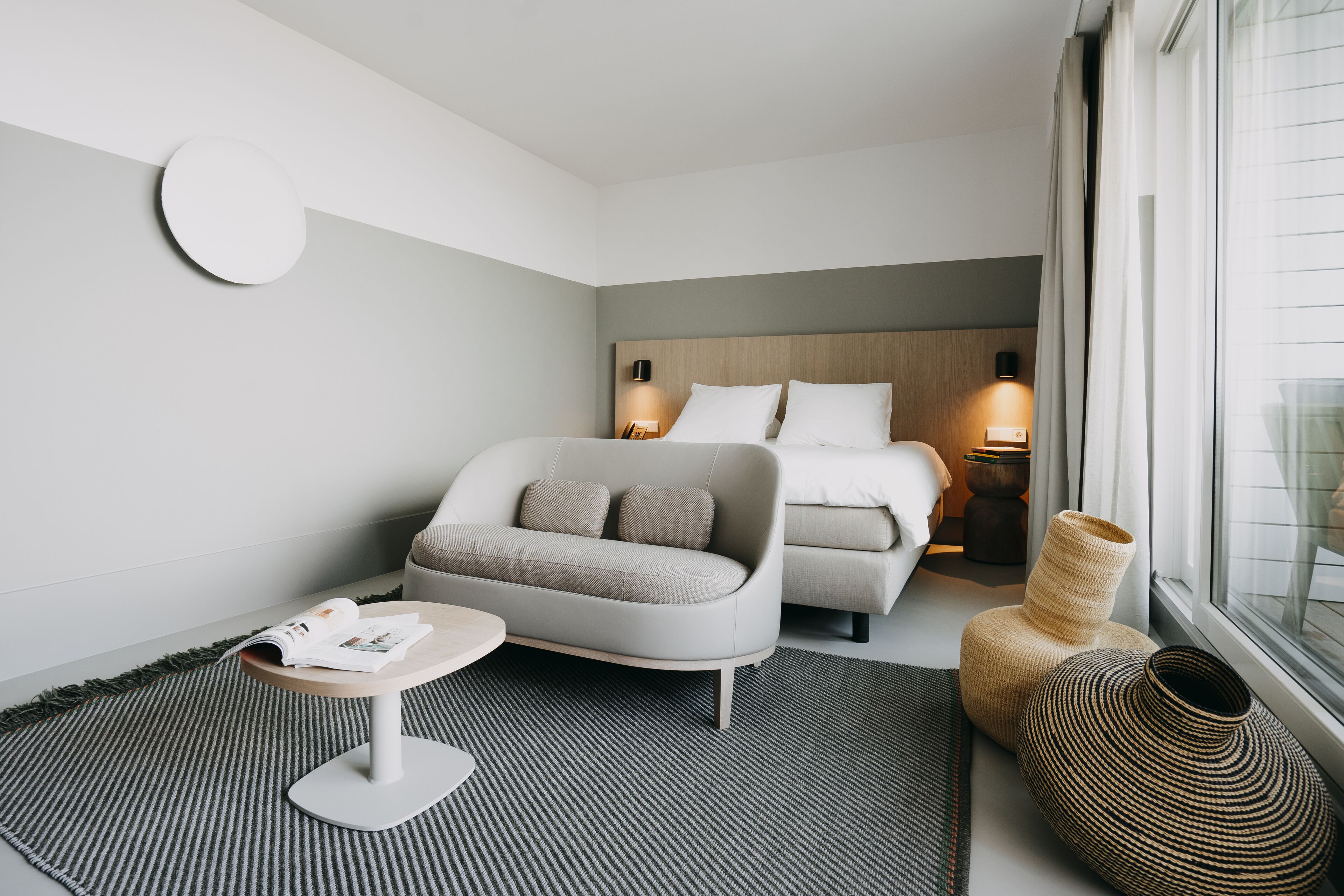 A modern hotel room with a bed, a sofa, a small table, and a large window.
