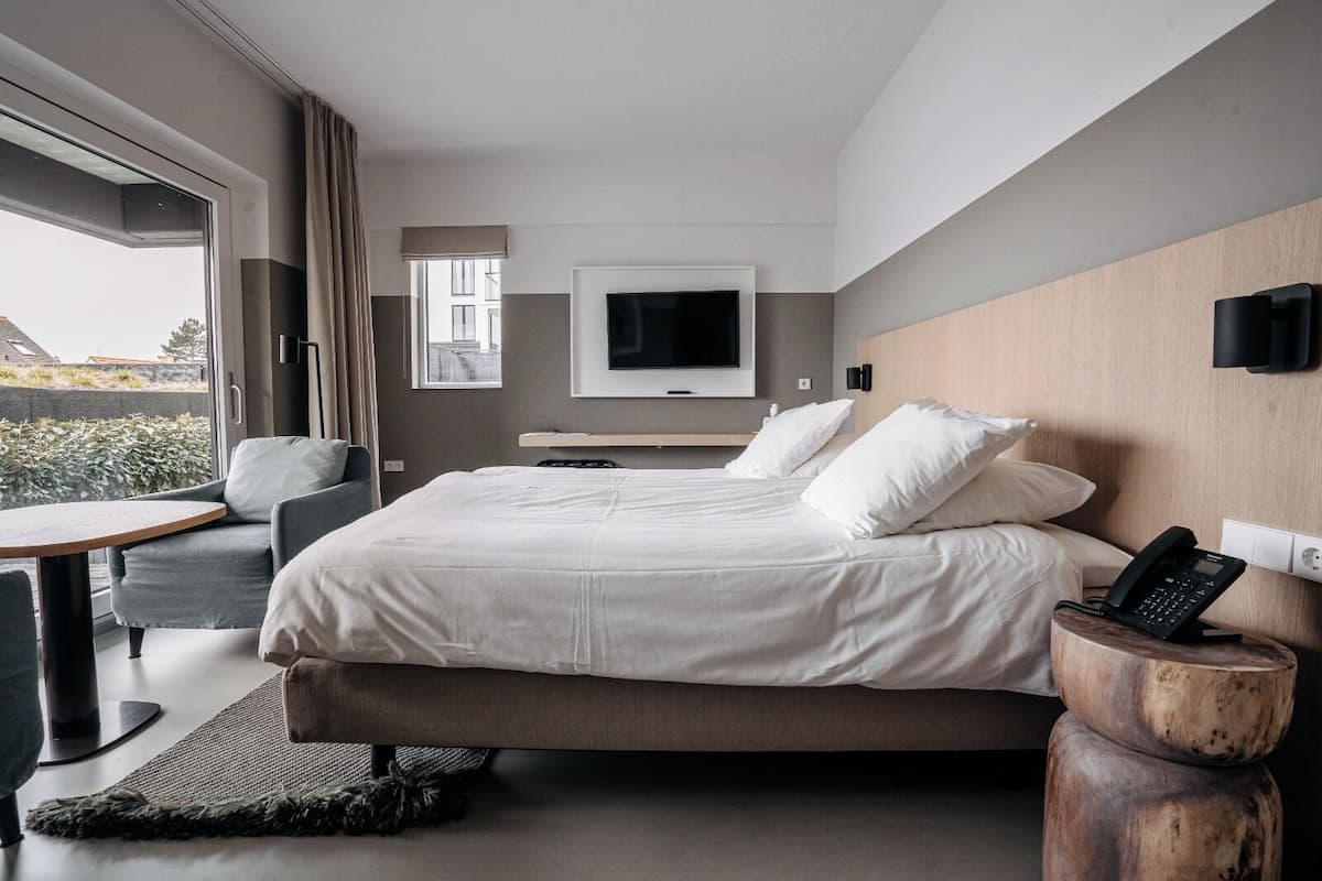 Una habitación de hotel moderna con una cama grande, un televisor montado en la pared, una mesita de noche de madera con un teléfono y un balcón con vistas a la ciudad.