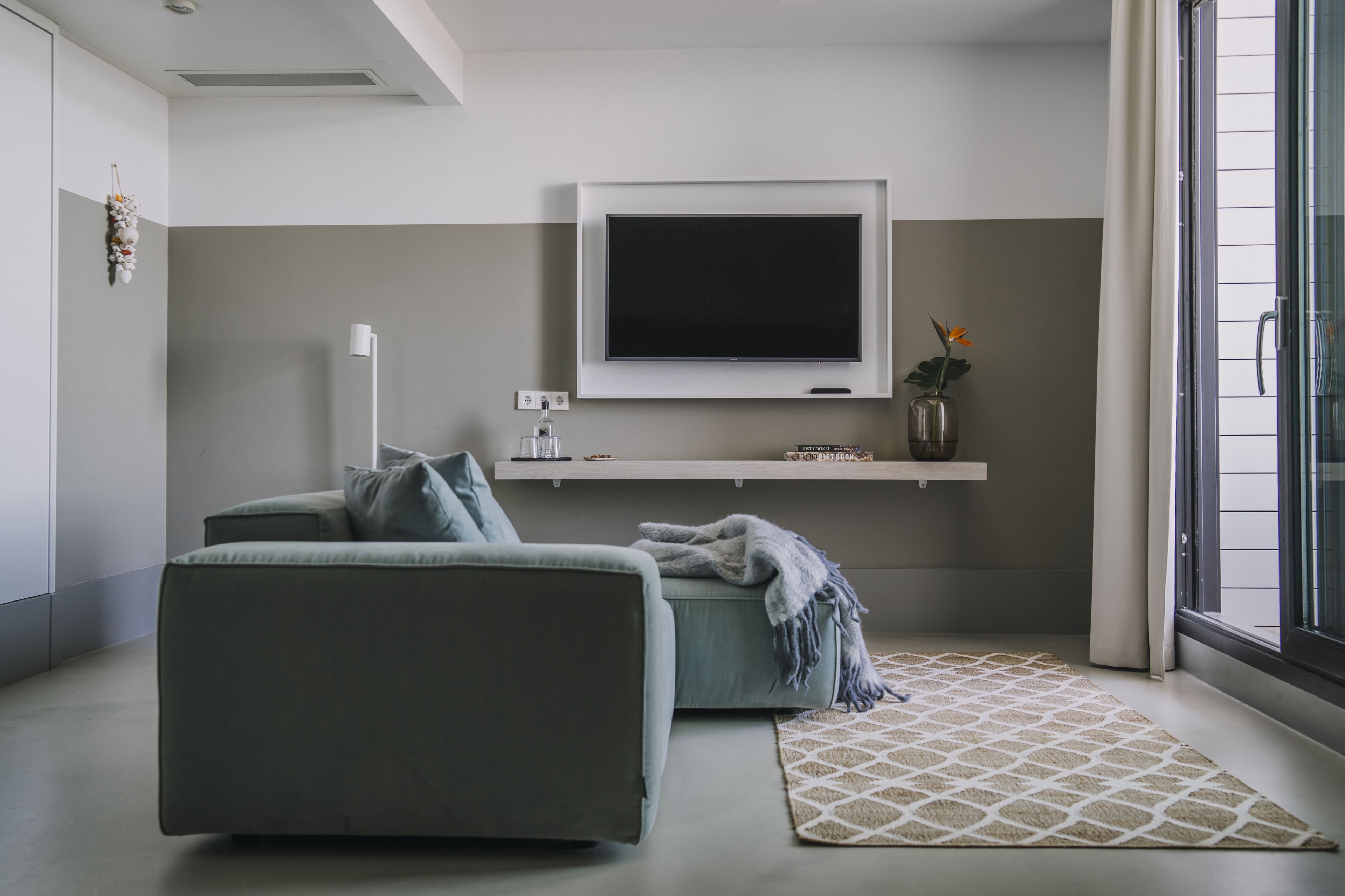 Een moderne woonkamer met een grijze bank, een flatscreen televisie aan de muur, een klein schap met decoratieve objecten, en een kamerkleed met dessin.