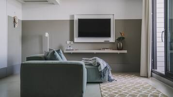 Een moderne woonkamer met een grijze bank, een flatscreen televisie aan de muur, een klein schap met decoratieve objecten, en een kamerkleed met dessin.