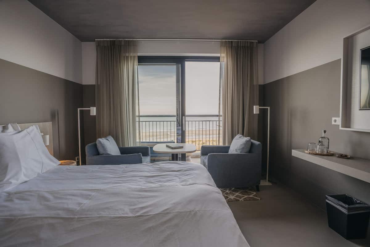 Habitación de hotel moderna con una cama grande, dos sillones, una mesita redonda y vistas amplias a través de un ventanal, además de una estantería con una botella y un vaso.