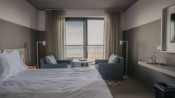 Een moderne hotelkamer met een groot bed, twee fauteuils, een klein rond tafeltje, uitzicht door een groot raam, en een plank met een fles en glas.
