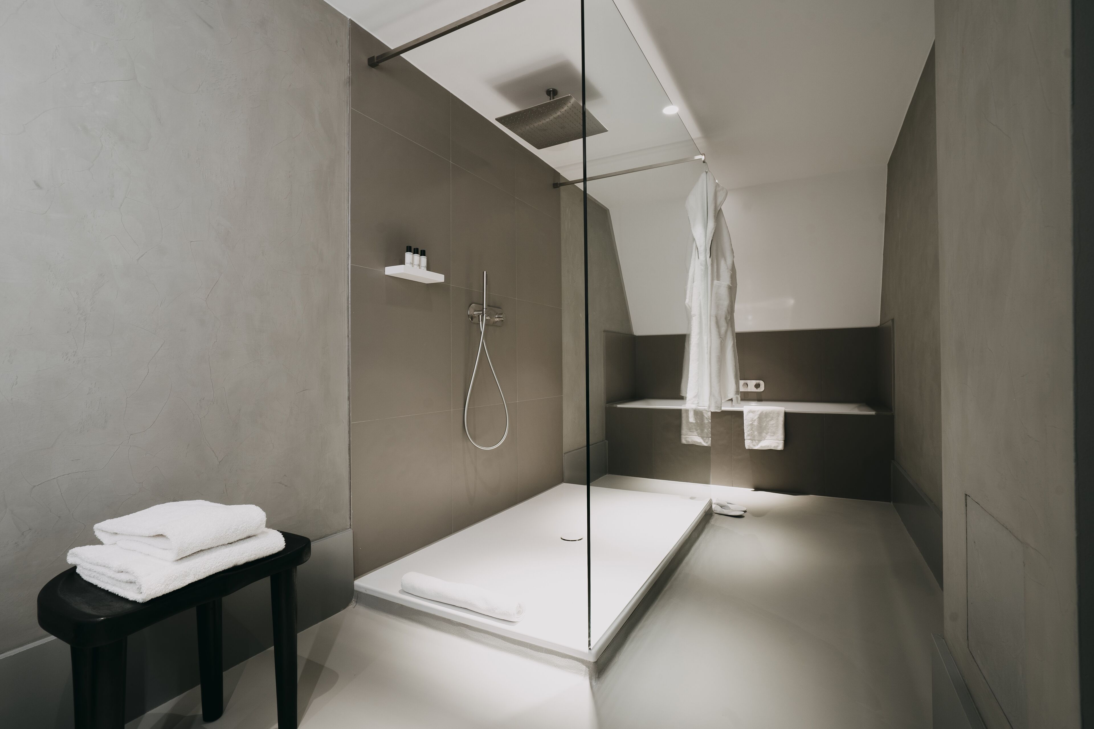 Een moderne badkamer met een douchecabine van glas, een zwart krukje met handdoeken en een minimalistisch design.