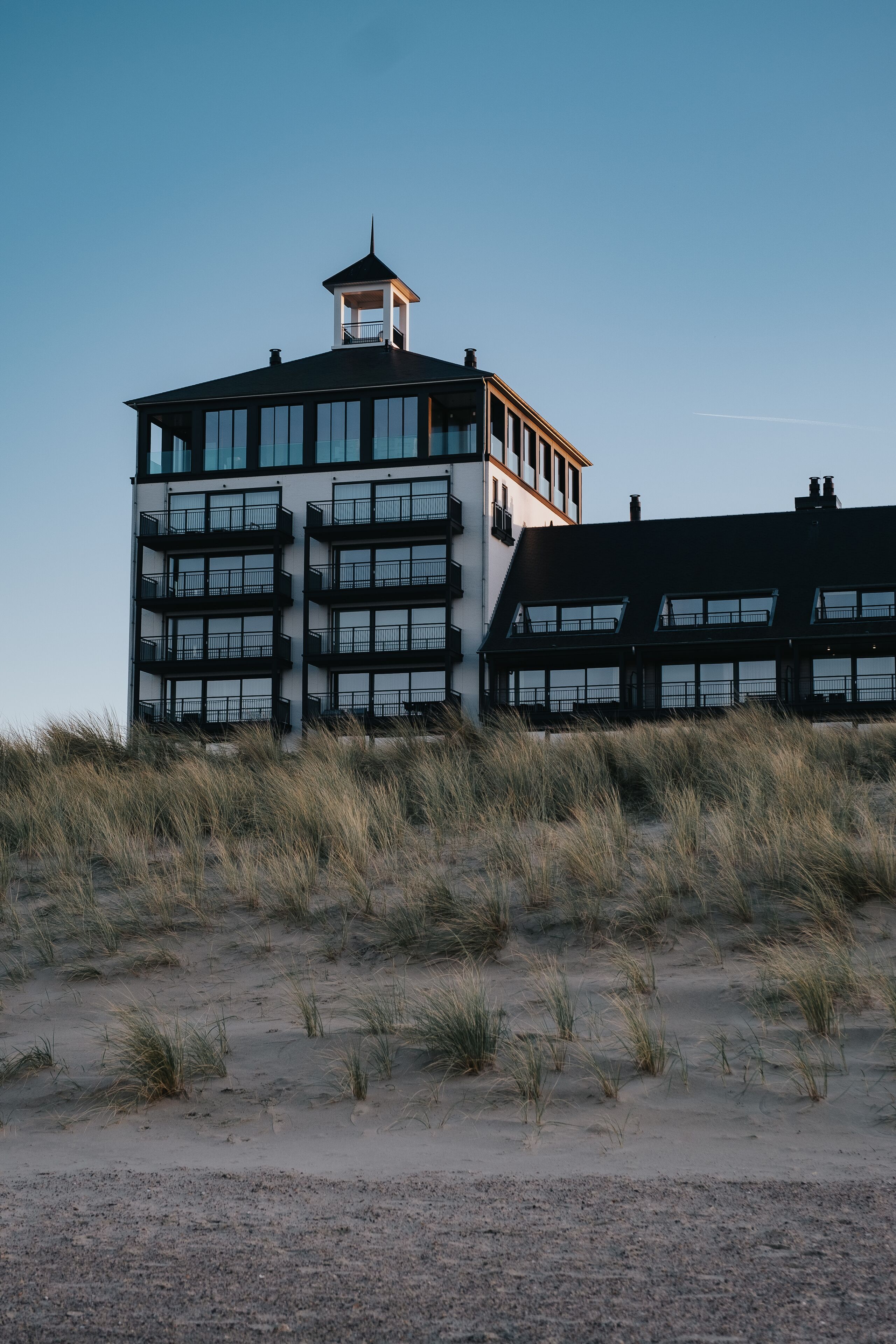 A modern building with multiple floors and a tower, surrounded by sand dunes.