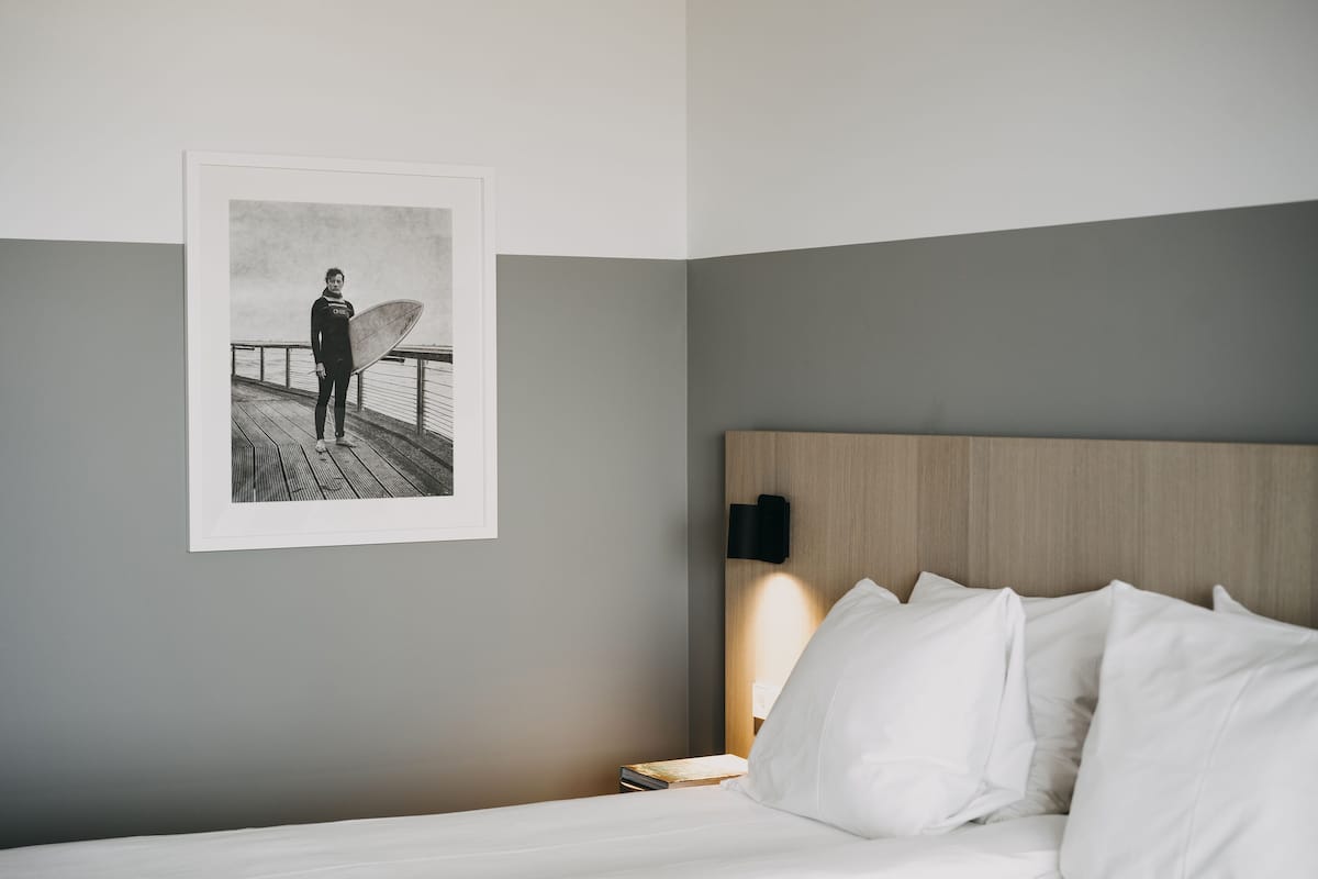 Habitación de hotel con cama, mesita de noche, un cuadro enmarcado de un surfista y una lámpara de pared.