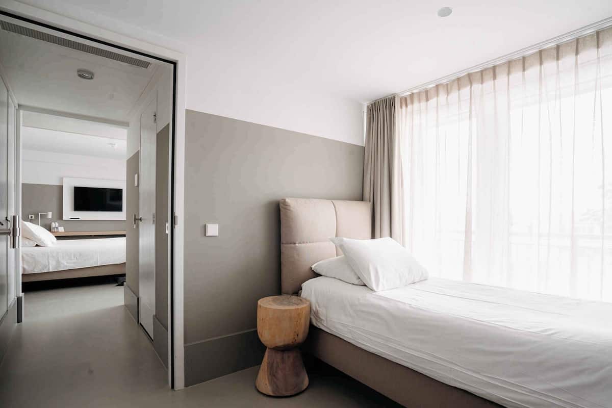 Una habitación de hotel moderna con una cama grande, un taburete de madera, un televisor y vistas a la ciudad a través de cortinas traslúcidas.