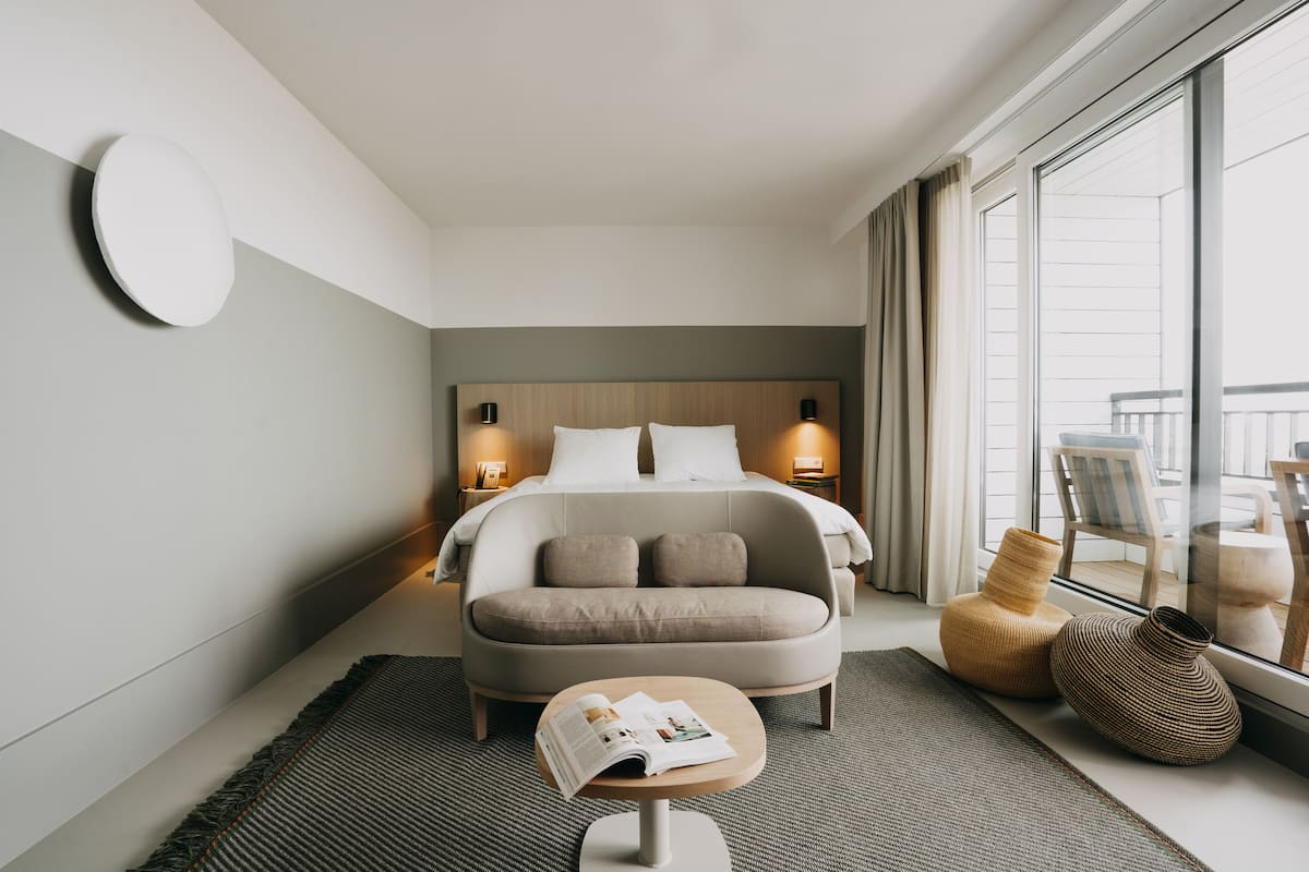 Una habitación de hotel moderna con una cama, lámparas de noche, una mesita con una revista y dos jarrones decorativos junto a la ventana.