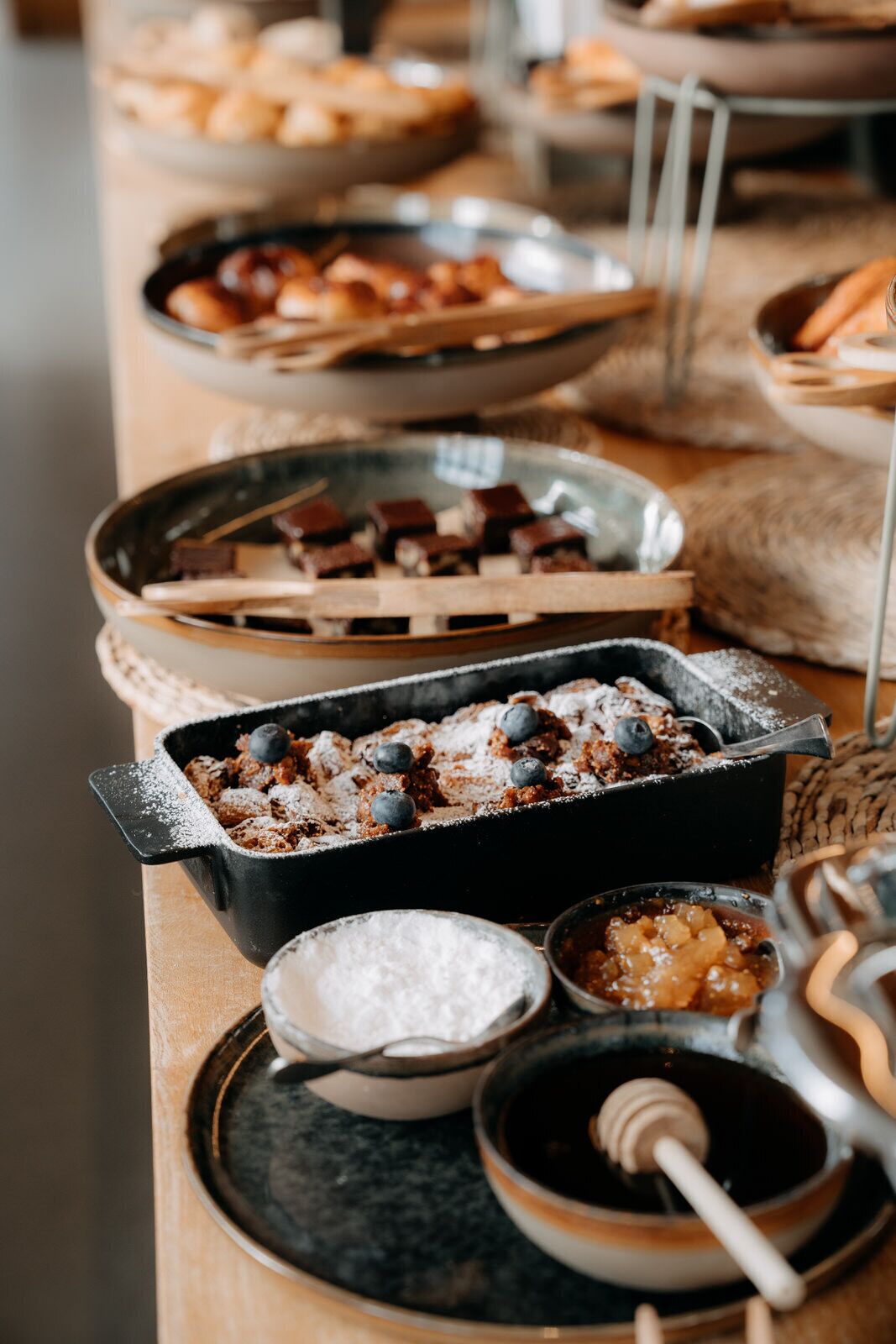 A buffet table with various dishes including a baked dessert with blueberries, a bowl of powdered sugar, and a honey dipper.