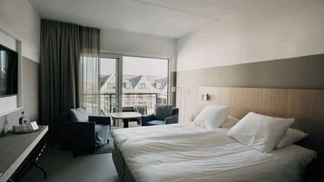 Een moderne hotelkamer met een groot bed, een bureau met stoel, een klein tafeltje en uitzicht op gebouwen door grote ramen.