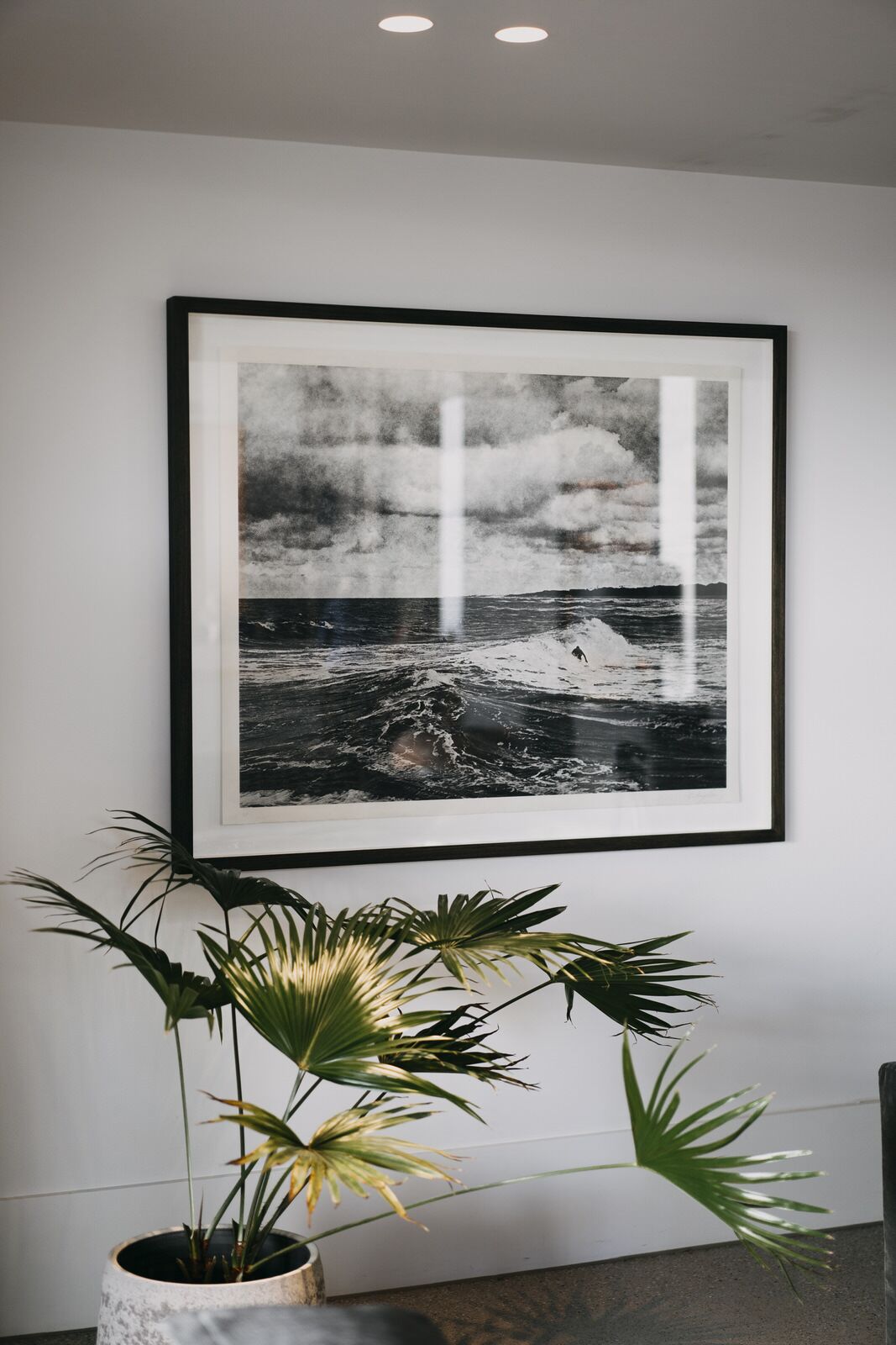 A framed black and white photograph of a stormy sea on a wall, with a potted plant in the foreground.