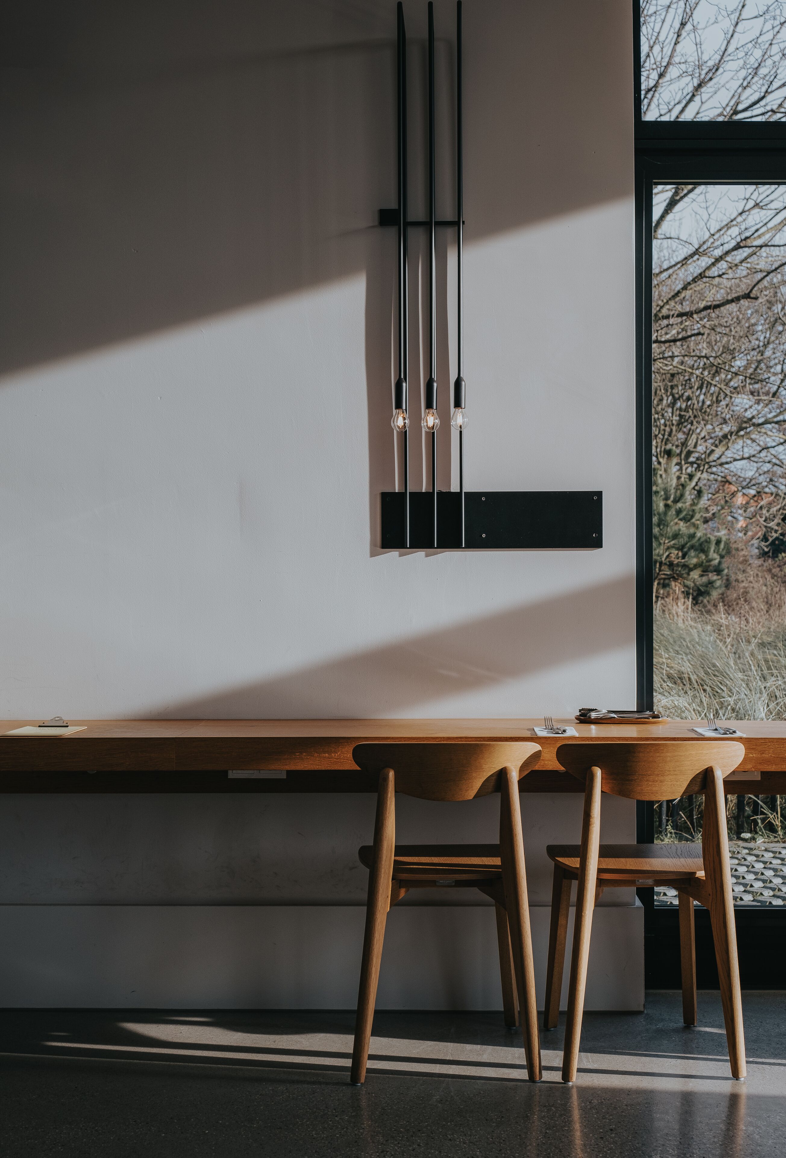 A modern dining area with a long wooden table and two chairs, a black wall-mounted light fixture, and a window with a view of trees.
