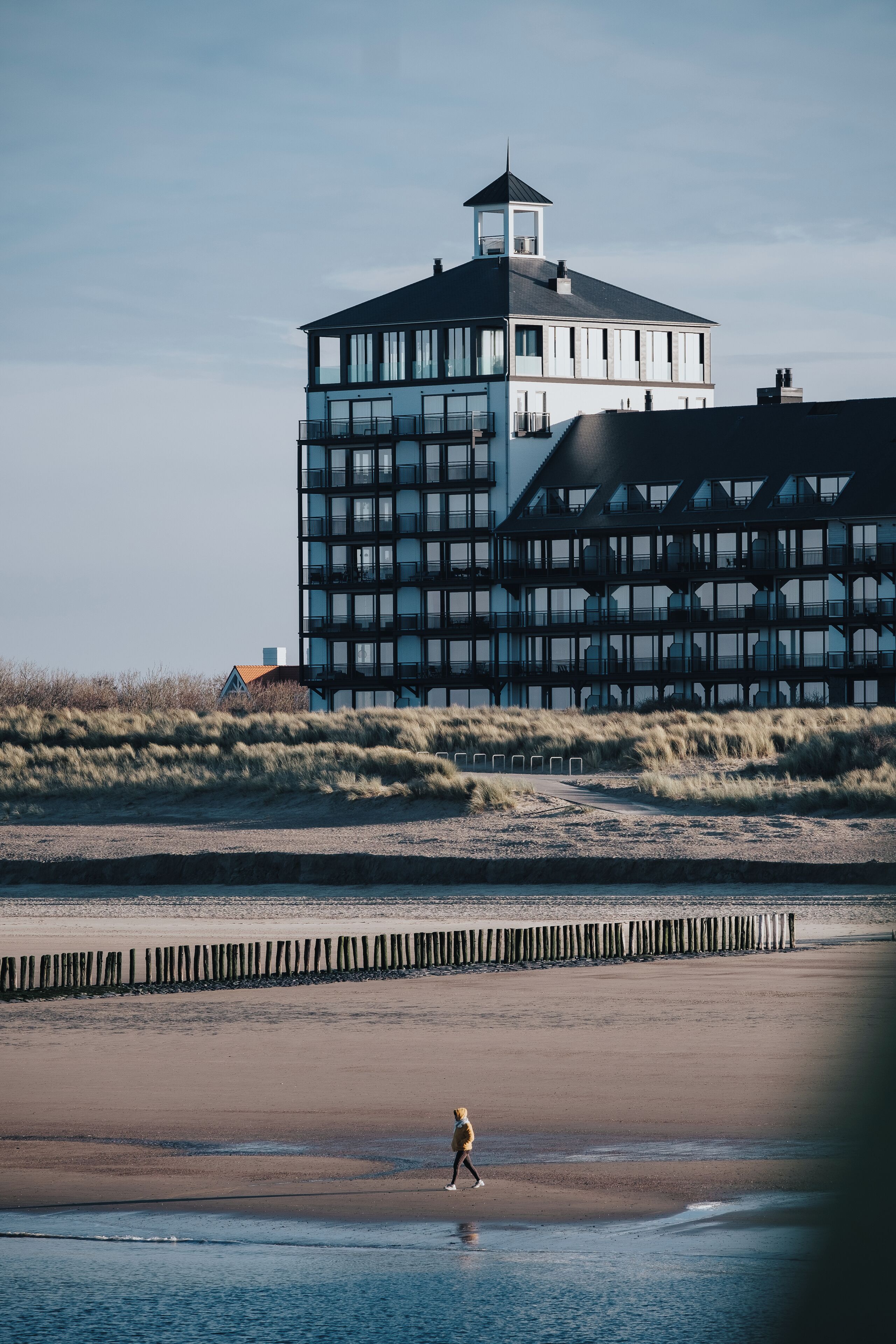 Een modern gebouw met meerdere verdiepingen en een toren, gelegen nabij een strand waar iemand over het zand loopt.