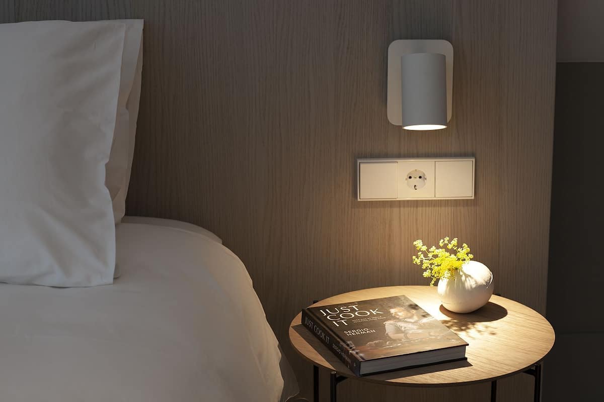 Una cama bien hecha con un almohadón blanco, una mesita de noche con un libro titulado 'Just Cook It' y un florero con flores amarillas, y una lámpara de pared sobre un enchufe.