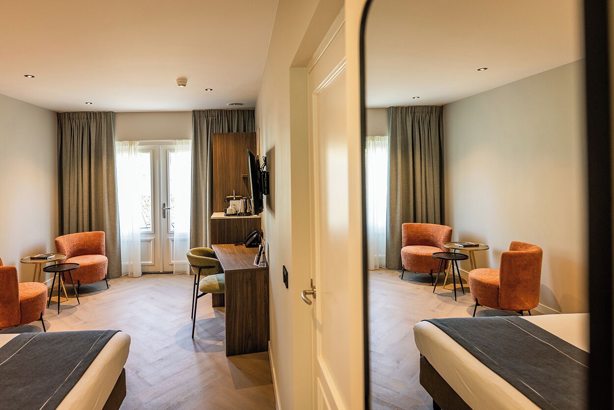 Ein Hotelzimmer mit einem Bett, zwei orangefarbenen Sesseln, einem kleinen Schreibtisch mit Stuhl und einem Spiegel, der den Raum reflektiert.