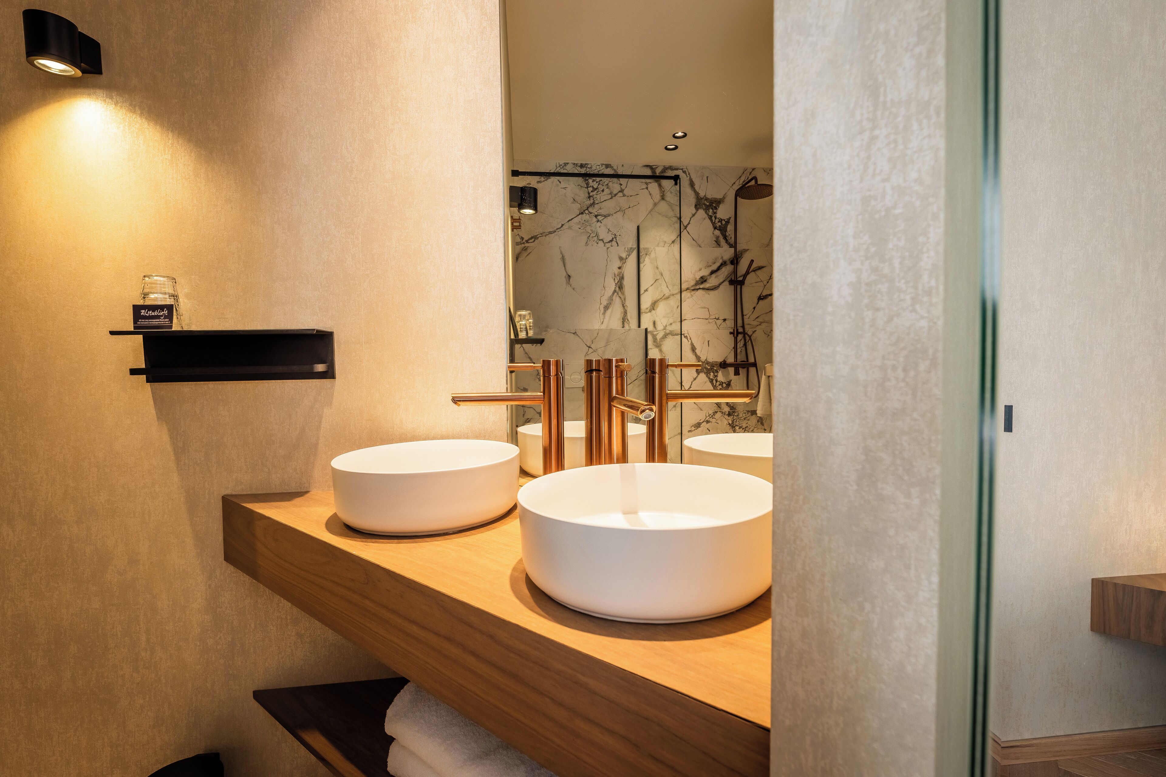 A modern bathroom with two round white sinks, wooden shelves, and a marble backsplash.