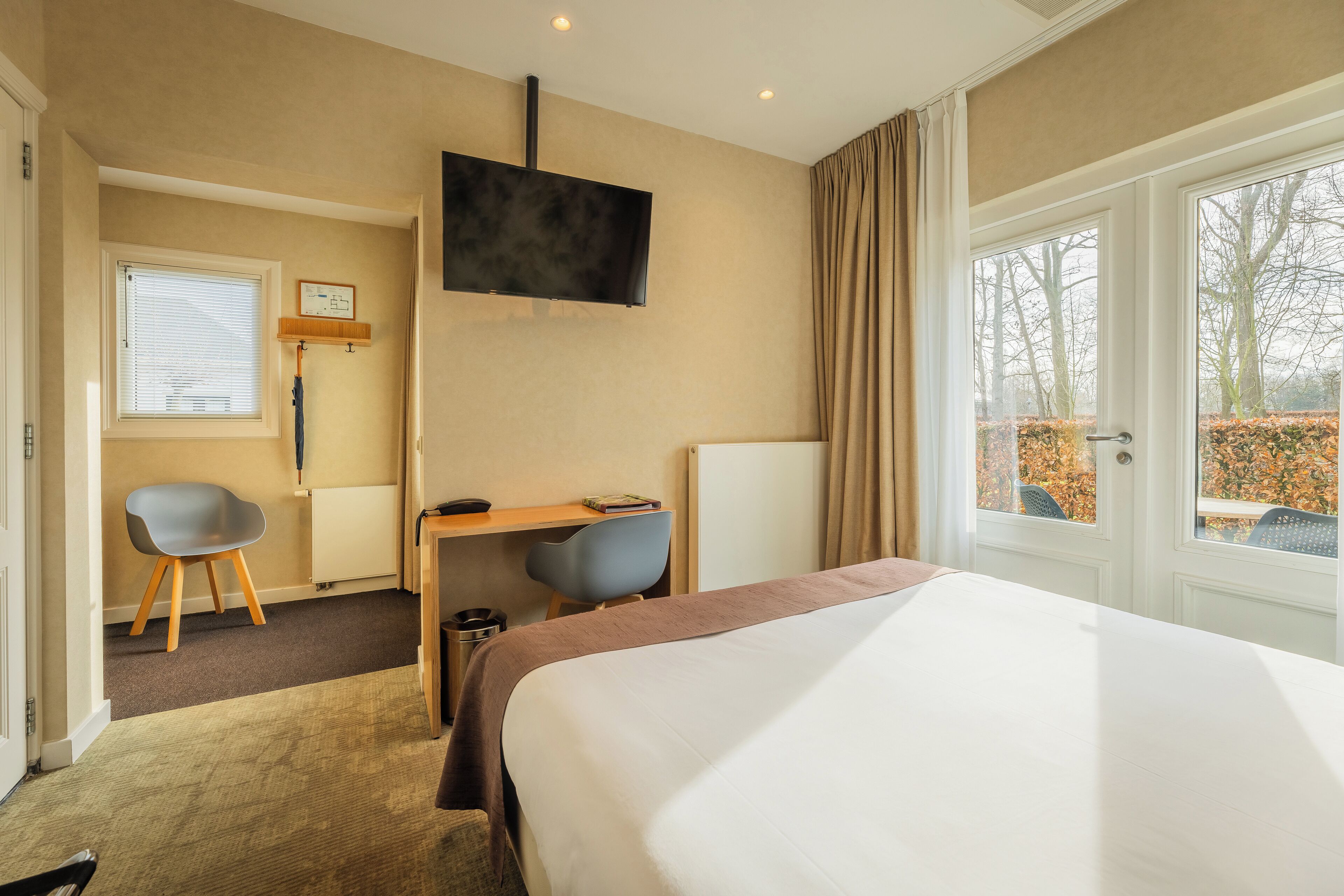A hotel room with a large bed, a desk with two chairs, a TV mounted on the wall, and a window with curtains.